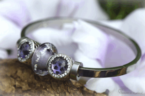 Dames bangle van RVS zilverkleurig met transparante paarse glasbedel met crematie-as en twee 925 zilveren bedels met paarse crystals, Silver-colored stainless steel women's bangle with transparent purple memorial glass bead containing ashes and two 925 silver charms with purple crystals, Silberfarbene Edelstahl-Damenarmband mit transparenter lila Glasperle mit Asche und zwei 925er Silber-Charms mit lila Kristallen